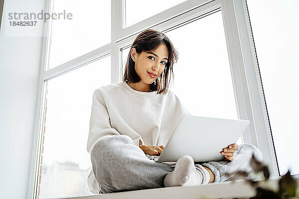 Lächelnde junge Frau sitzt mit Laptop vor dem Fenster zu Hause