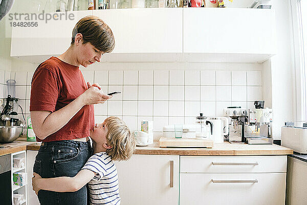 Junge umarmt Mutter mit Smartphone  das in der Küche steht