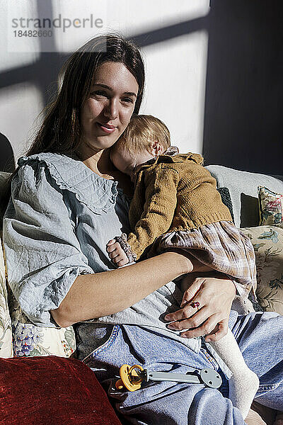 Lächelnde Mutter mit kleinem Mädchen  das im Sonnenlicht auf dem Sofa sitzt