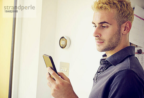 Junger Mann benutzt Smartphone und stellt zu Hause Thermostat ein