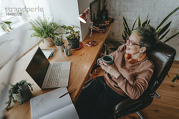 Lächelnde Geschäftsfrau mit Kaffeetasse sitzt im Heimbüro