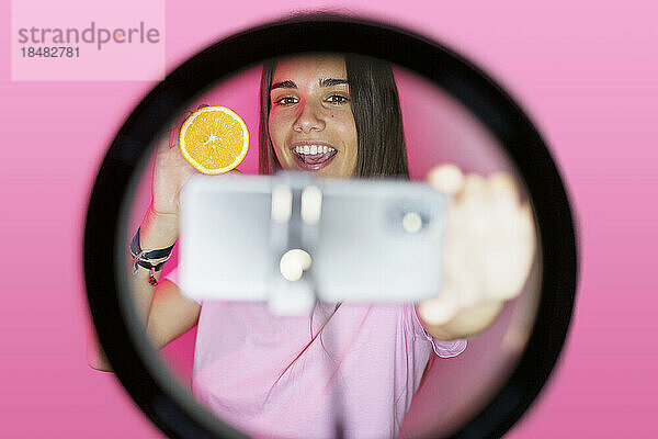 Lächelnder Influencer filmt über Orange auf Smartphone im Ringlicht