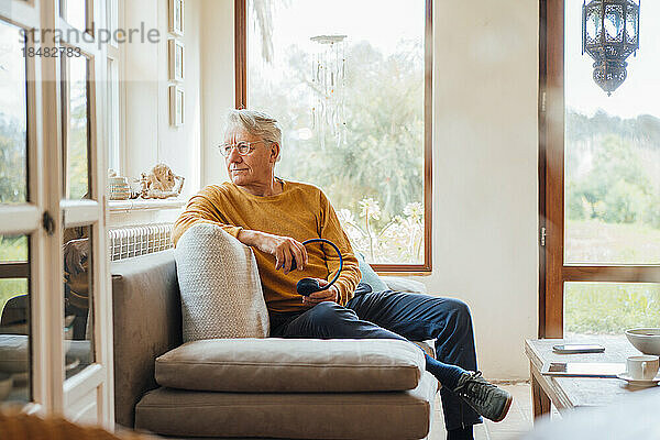 Nachdenklicher älterer Mann mit kabellosen Kopfhörern sitzt zu Hause auf dem Sofa