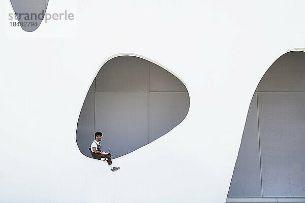 Junger Mann mit Laptop sitzt an der Wand