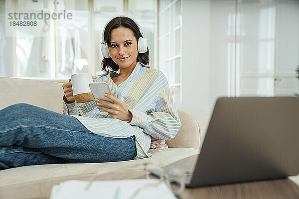 Lächelnder Freiberufler mit kabellosen Kopfhörern sitzt mit Smartphone und Kaffeetasse auf dem heimischen Sofa