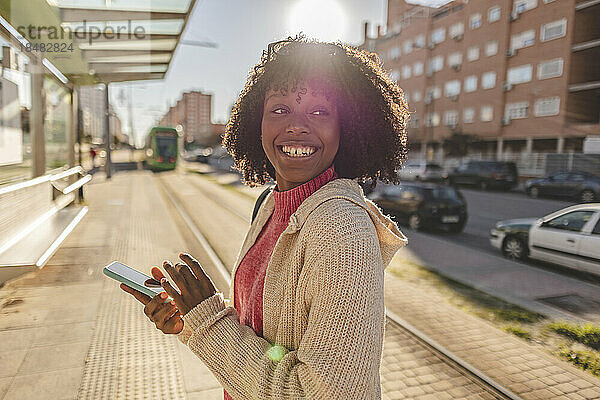Glückliche junge Frau mit Smartphone steht am Bahnhof