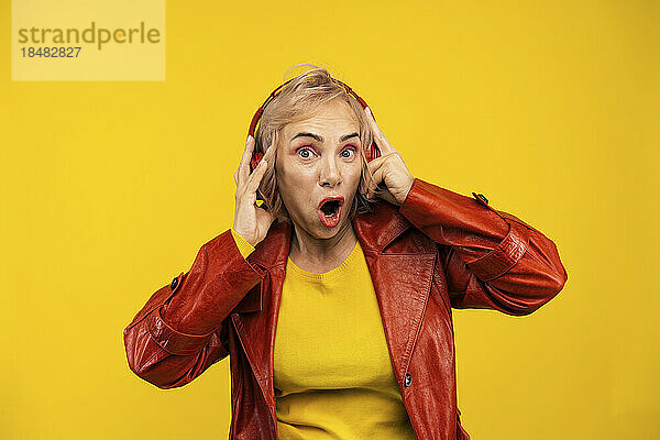 Schockierte ältere Frau mit kabellosen Kopfhörern  die vor gelbem Hintergrund Musik hört