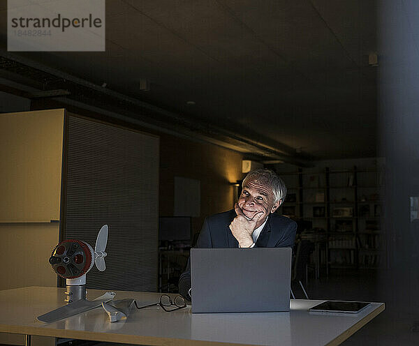 Lächelnder Geschäftsmann mit der Hand am Kinn sitzt am Schreibtisch im Büro