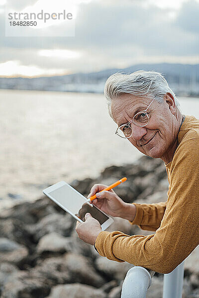 Lächelnder älterer Mann mit Tablet-PC und digitalisiertem Stift lehnt am Geländer