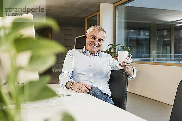 Glücklicher älterer Geschäftsmann mit Tasse sitzt am Schreibtisch im Büro