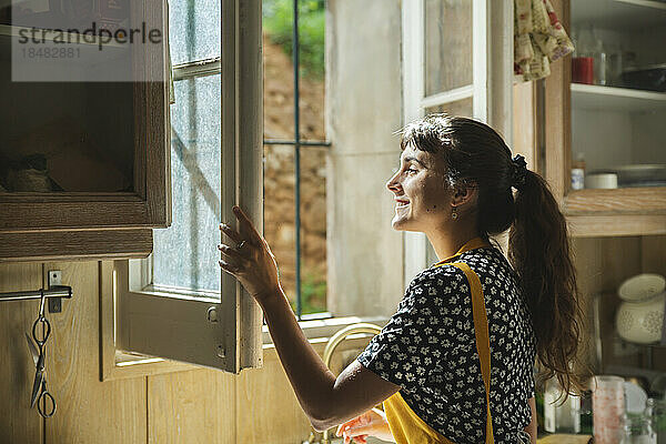 Lächelnde junge Frau  die durch das Fenster in der Küche schaut