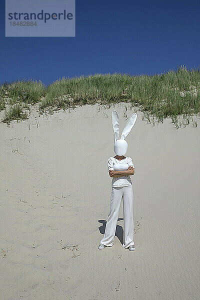 Woman wearing rabbit mask standing with arms crossed on sand dune