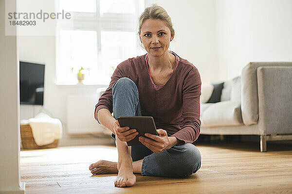 Frau hält Tablet-PC und sitzt zu Hause auf dem Boden