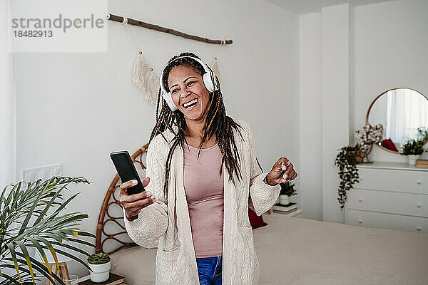 Glückliche Frau mit kabellosen Kopfhörern steht im Schlafzimmer