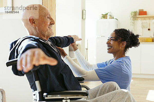 Lächelnder Physiotherapeut untersucht älteren Mann  der zu Hause die Arme ausstreckt