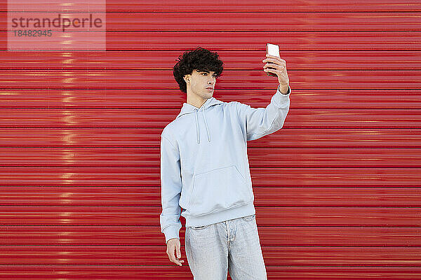 Junger Mann macht Selfie mit Smartphone vor roter Wand