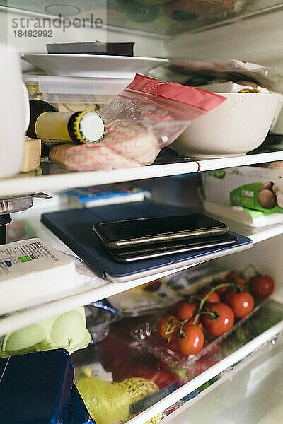 Smartphone und Tablet-PC mit gestapelten Lebensmitteln auf dem Regal im Kühlschrank
