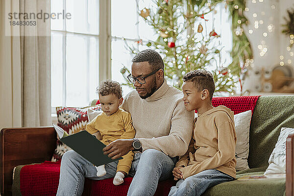 Vater mit Tochter und Sohn lesen zu Weihnachten ein Buch auf dem Sofa