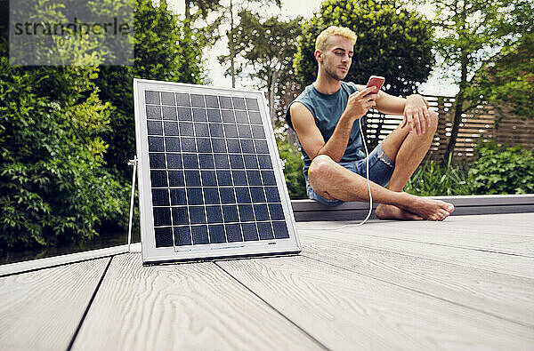 Junger Mann sitzt auf der Terrasse und nutzt sein Smartphone  das über ein Solarpanel aufgeladen wird