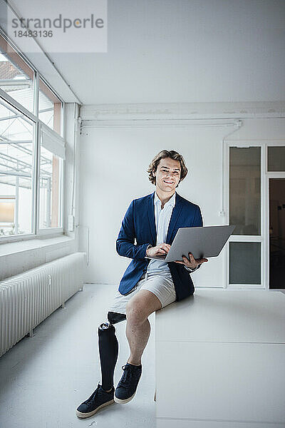 Glücklicher Geschäftsmann mit Prothesenausrüstung  der mit Laptop auf dem Schreibtisch sitzt