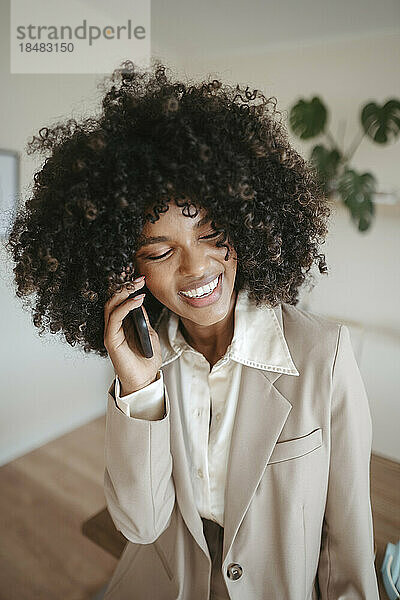 Glückliche Geschäftsfrau mit Afro-Frisur  die im Büro auf dem Smartphone spricht