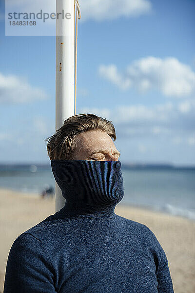 Mann bedeckt Gesicht mit Rollkragenpullover und entspannt sich am Strand