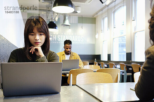 Junge Geschäftsfrau mit der Hand am Kinn und Laptop in der Cafeteria