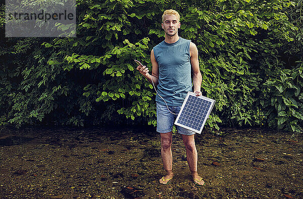 Junger Mann steht im Bach und hält sein Mobiltelefon in der Hand  das über ein Solarpanel aufgeladen wird