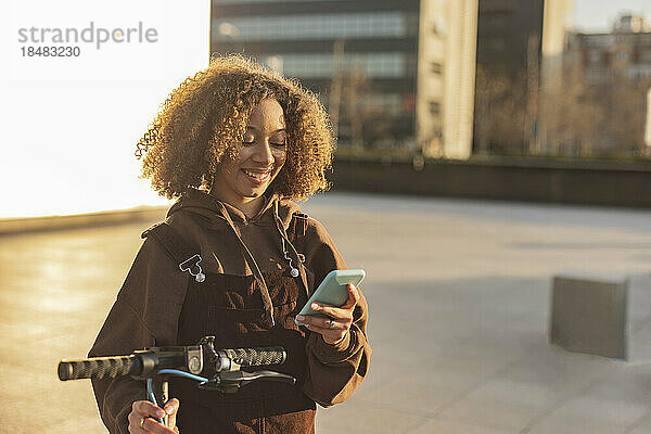 Lächelnde Frau mit Tretroller und Mobiltelefon bei Sonnenuntergang