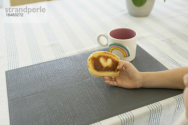 Hand of girl holding pancake with heart shape by mug on table at home