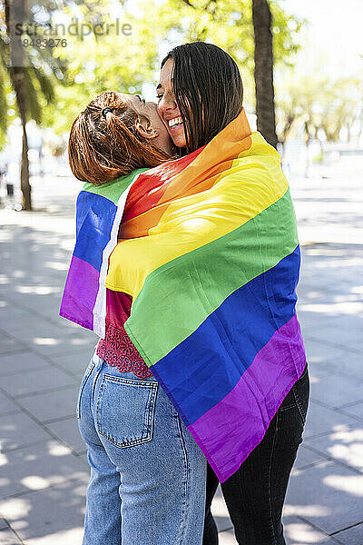 Lesbisches Paar umarmt sich in regenbogenfarbener Flagge auf Fußweg
