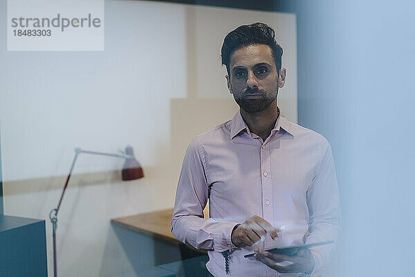 Contemplative businessman with tablet PC standing in office seen through glass