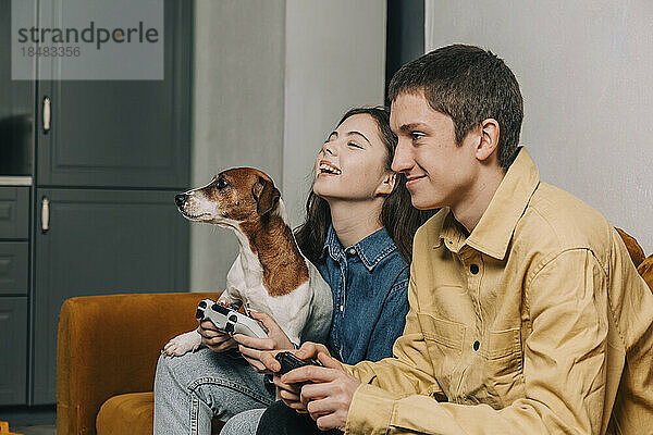 Glückliches Teenager-Paar mit Hund spielt zu Hause auf dem Sofa