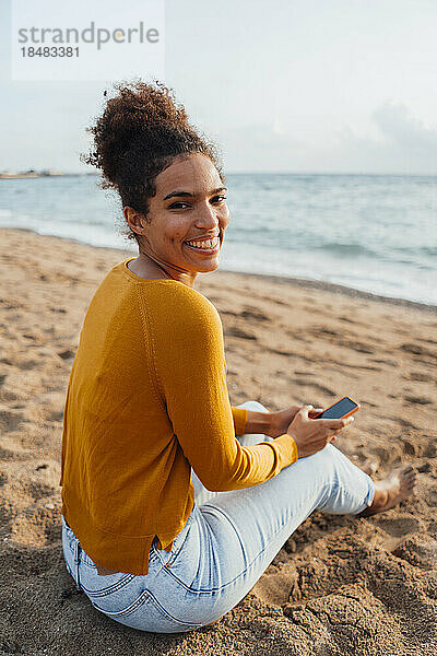 Glückliche junge Frau sitzt mit Handy am Strand