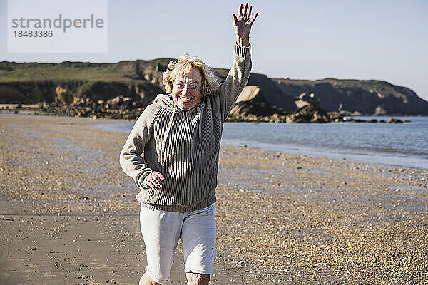 Cheerful senior woman with arm raised running at beach