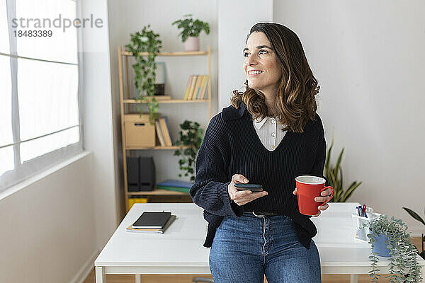 Nachdenkliche Geschäftsfrau lehnt mit Smartphone und Kaffeetasse am Schreibtisch