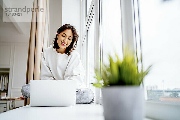 Smiling young woman using laptop on window sill