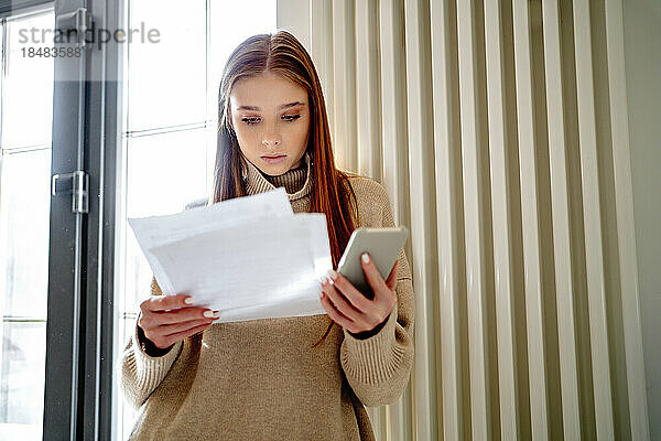Teenage girl holding energy bill and mobile phone by radiator heater at home