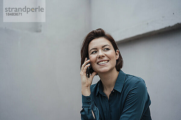 Glückliche Geschäftsfrau  die mit ihrem Smartphone an einer grauen Wand spricht