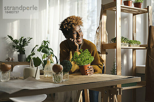 Glückliche Frau mit Topfpflanze  die zu Hause am Tisch lehnt