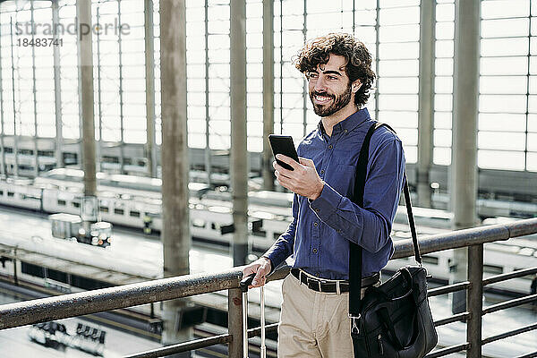 Glücklicher Geschäftsmann mit Smartphone  der auf der Brücke am Bahnhof steht