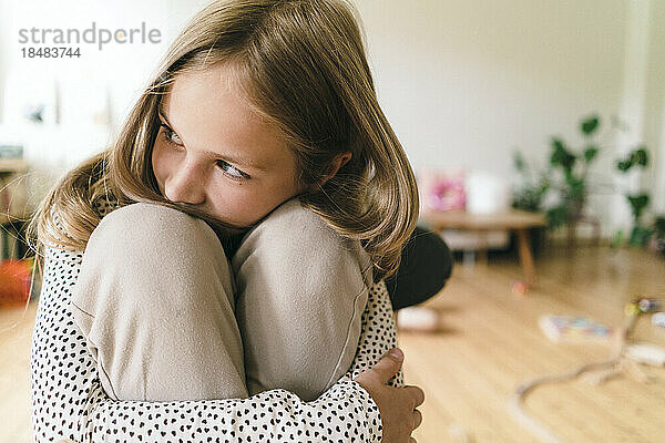 Nachdenkliches Mädchen umarmt Knie  die zu Hause sitzen