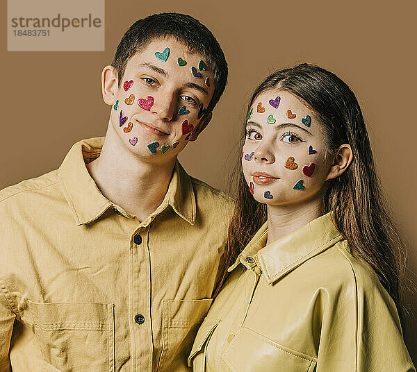 Lächelndes Teenager-Paar mit herzförmigen Aufklebern im Gesicht vor der Wand