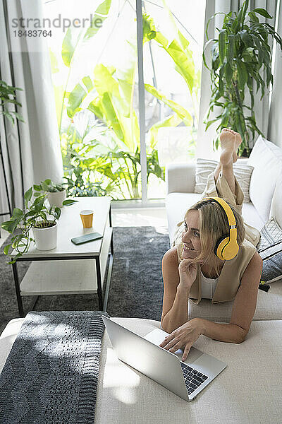 Glücklicher Freiberufler mit kabellosen Kopfhörern  der mit Laptop auf dem Sofa liegt