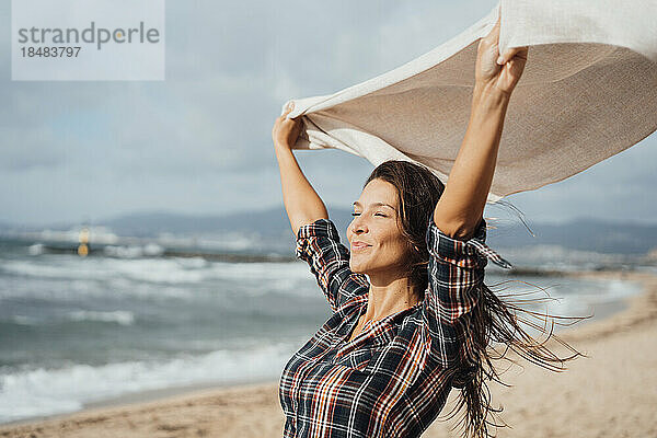 Lächelnde Frau mit Schal und erhobenen Armen am Strand