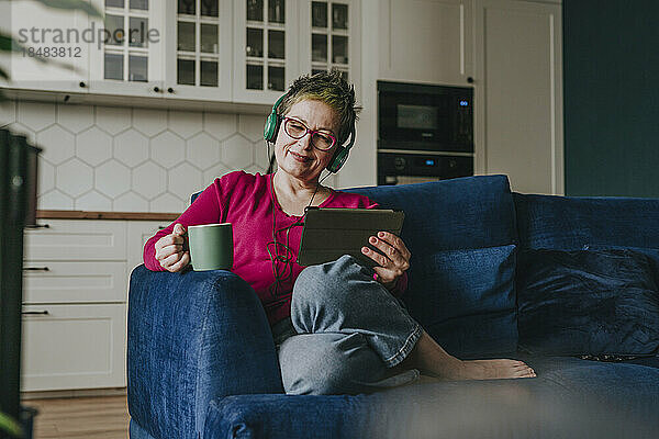 Lächelnde reife Frau mit Kopfhörern und Kaffeetasse auf dem heimischen Sofa