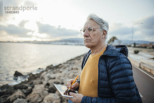 Älterer Mann steht mit Tablet-PC vor dem Meer an der Promenade