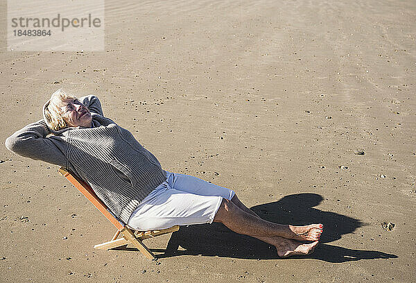 Ältere Frau mit den Händen hinter dem Kopf entspannt sich an einem sonnigen Tag am Strand