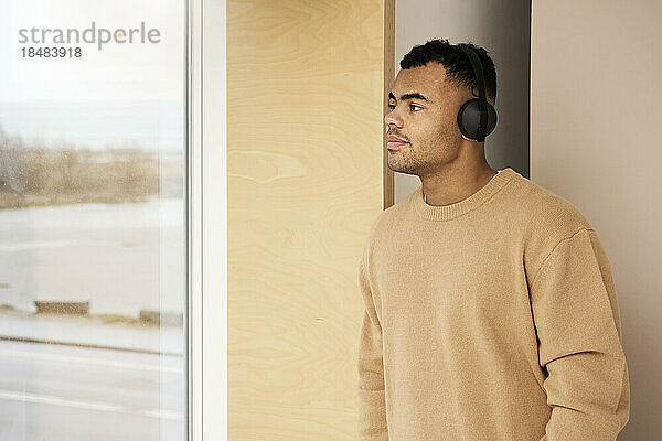 Young man looking out of window wearing wireless headphones