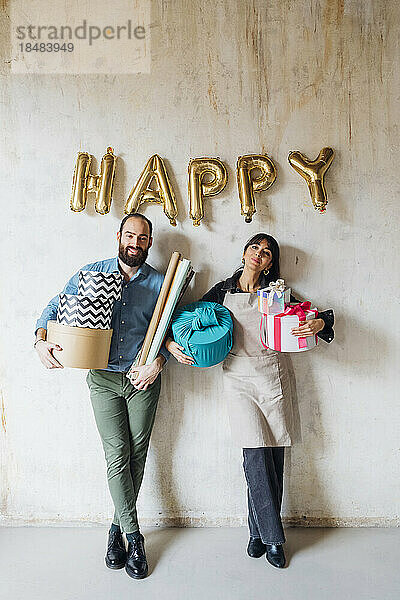 Smiling couple holding gift boxes leaning on wall